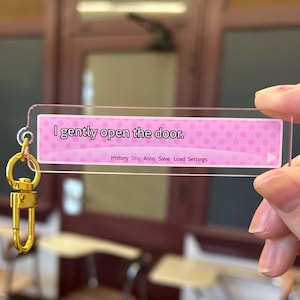 Puede incluir: Un llavero acrílico transparente con una inserción rectangular rosa. La inserción tiene el texto "I gently open the door." y un patrón de puntos. Un cierre y un anillo dorados están unidos al llavero.
