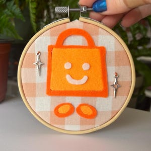 May include: Orange felt bag with a smiling face, two orange felt circles, and two silver star-shaped earrings on a white and orange gingham fabric background.
