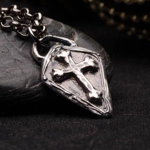May include: A silver pendant necklace with a cross design. The cross is set within a shield-shaped charm. The pendant is on a silver chain.