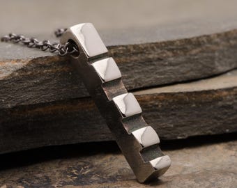 May include: A silver-toned pendant necklace featuring a rectangular bar with a series of square, raised elements. The pendant hangs from a dark chain. The jewelry is displayed on a textured, gray stone surface.