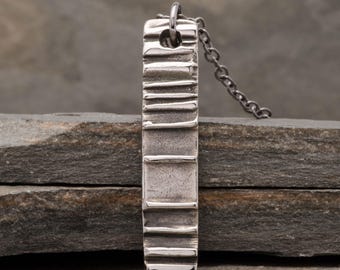 May include: A silver pendant necklace with a rectangular, textured pendant. The pendant has horizontal lines and a small hole at the top for the chain. The chain is a dark silver color.