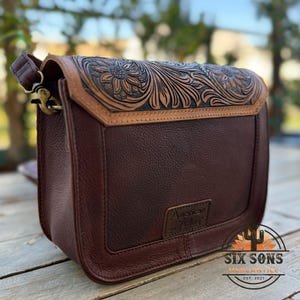 May include: A brown leather crossbody bag with a floral embossed design on the flap. The bag features a dark brown body with a lighter brown trim and strap. The words "American Darling" are embossed on the front.
