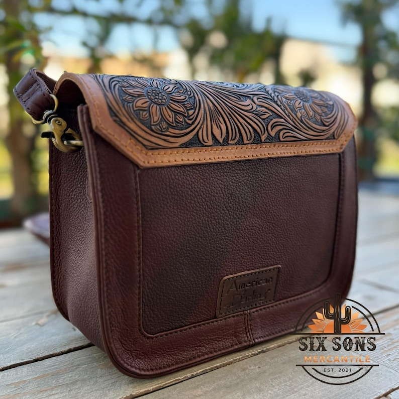 May include: A brown leather crossbody bag with a floral embossed design on the flap. The bag features a dark brown body with a lighter brown trim and strap. The words "American Darling" are embossed on the front.