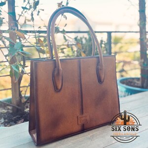 Puede incluir: Un bolso de mano estructurado de cuero marr&oacute;n con dos asas superiores y una costura vertical en el centro. Un peque&ntilde;o parche rectangular con el texto American Darling en relieve se encuentra en la parte inferior. El bolso tiene un acabado mate.