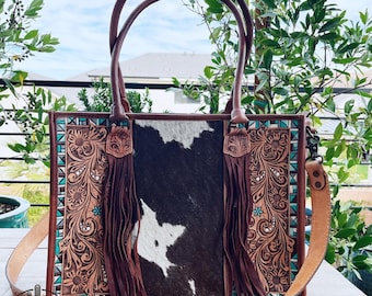 May include: A rectangular handbag featuring a central panel of black and white cowhide leather flanked by two panels of tan leather with intricate floral tooling and turquoise accents. The bag includes long brown leather fringe and a shoulder strap.