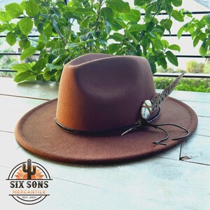 May include: A brown felt fedora hat with a black leather band and a feather accent. The hat has a white stone and a brown leather cord attached to the feather.
