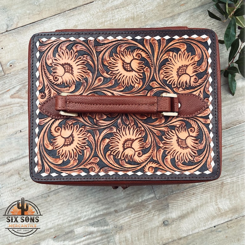 May include: A brown leather box with a tooled floral design and a brown leather handle. The box has a white border with a diamond pattern.