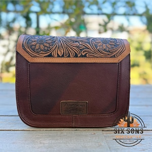 May include: A brown leather crossbody bag with a floral embossed design on the flap. The bag features a rectangular shape with a stitched border and a small rectangular tag that reads "American Darling". The bag has a brown leather strap.