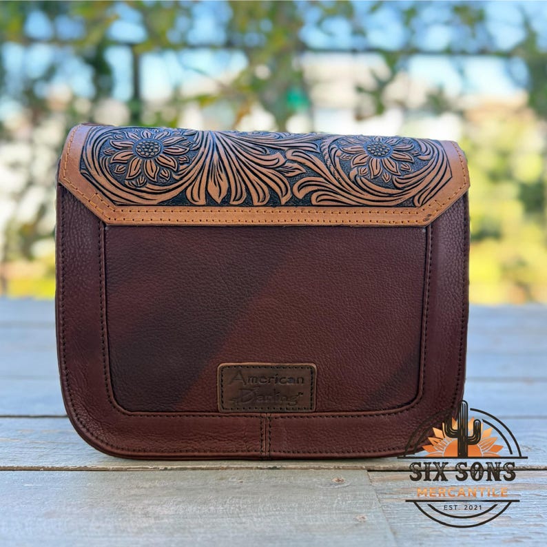 May include: A brown leather crossbody bag with a floral embossed design on the flap. The bag features a rectangular shape with a stitched border and a small rectangular tag that reads "American Darling". The bag has a brown leather strap.