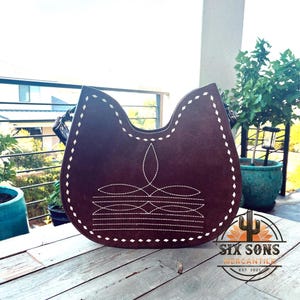 May include: A brown suede handbag with a unique shape and white stitching. The bag features a decorative stitched design and a handle. The bag is sitting on a wooden surface.