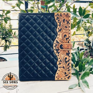 May include: A black leather folder with a quilted design and a decorative floral tooled leather border. The border features a sunflower design and intricate floral patterns.