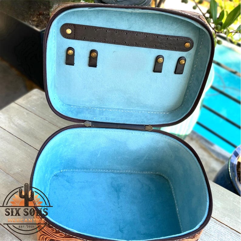 May include: Open, oval-shaped jewelry case with a brown exterior featuring a detailed embossed pattern. The interior is lined with light blue fabric. The case has a brown leatherette top with gold-colored hooks for hanging jewelry.