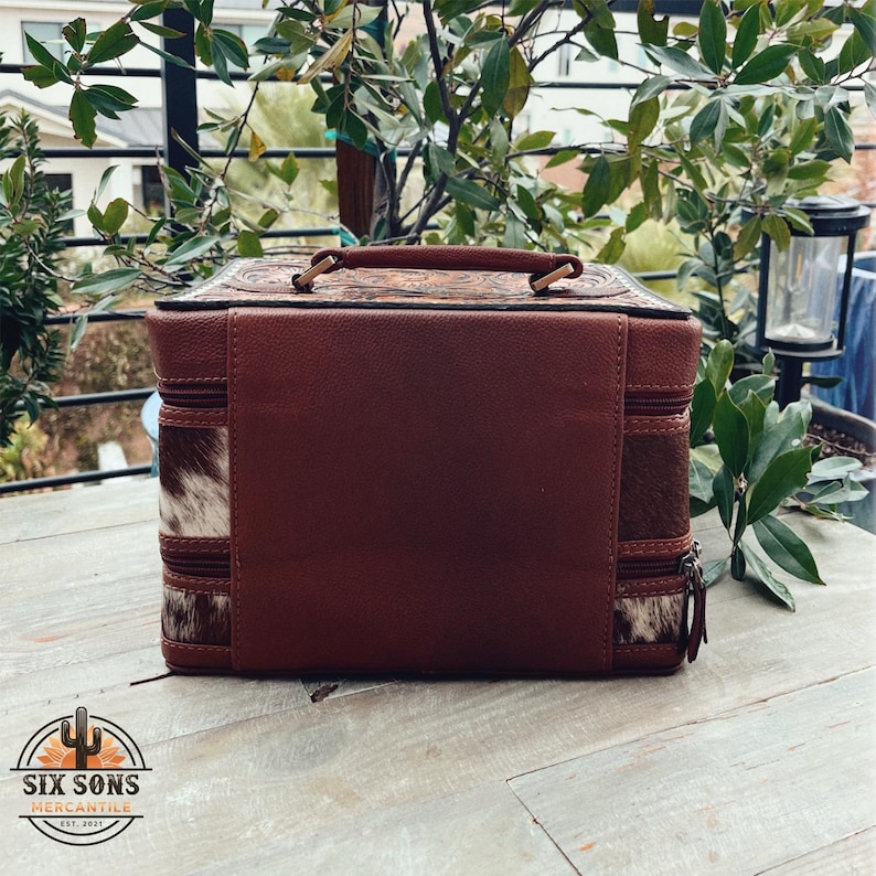 May include: A brown leather and cowhide travel case with a zipper closure. The case has a decorative floral design on the top and a brown leather handle.