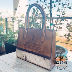 Puede incluir: Un bolso de mano estructurado de cuero marr&oacute;n con dos asas, un patr&oacute;n de costuras decorativo en la parte superior frontal y un panel de piel de vaca texturizado en blanco y negro en la base, separado por un borde de cuero trenzado oscuro.