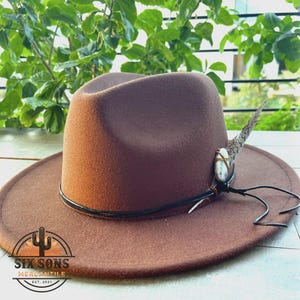 May include: A brown fedora hat with a black leather band and a small white stone and feather accent. The hat is sitting on a wooden surface. The hat has a logo on the brim that says "Six Sons Mercantile Est. 2021".