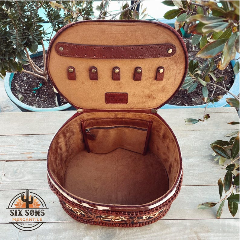 May include: A brown leather round hat box with a white and brown cowhide pattern. The lid is open and the interior is lined with brown fabric. There are five small compartments on the lid for storing jewelry.