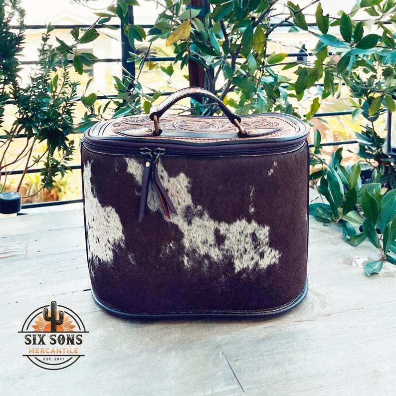 May include: A brown leather oval-shaped travel case with a white and brown cowhide pattern. The case has a zipper closure and a top handle.