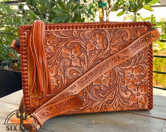 May include: A brown leather crossbody bag with a floral embossed design. The bag features a long, adjustable strap and a decorative fringe tassel. The bag has a rectangular shape and is made of a light brown leather.