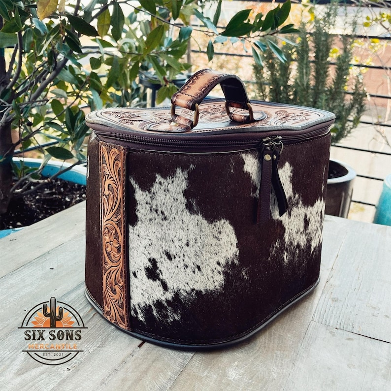 May include: A brown and white cowhide leather travel bag with a zipper closure and a brown leather handle. The bag has a floral tooled design on the side.