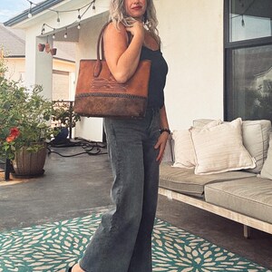 Puede incluir: Un bolso tote de cuero estructurado con un dise&ntilde;o bicolor que presenta una secci&oacute;n superior en color tostado claro y un panel inferior texturizado en marr&oacute;n oscuro. Incluye patrones de costuras blancas y un detalle de borde de cuero trenzado.