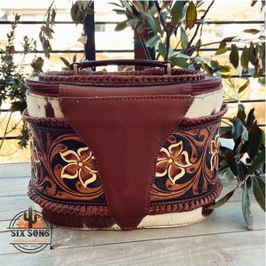May include: A brown leather and cowhide travel bag with a floral design and a braided handle. The bag has a zipper closure and a brown leather flap with a decorative stitching detail. The bag is labeled "Six Sons Mercantile Est. 2021".