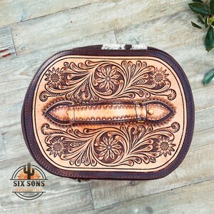 May include: A brown leather oval-shaped bag with a tooled floral design and a brown leather handle. The bag has a zipper closure and is made of high-quality leather.