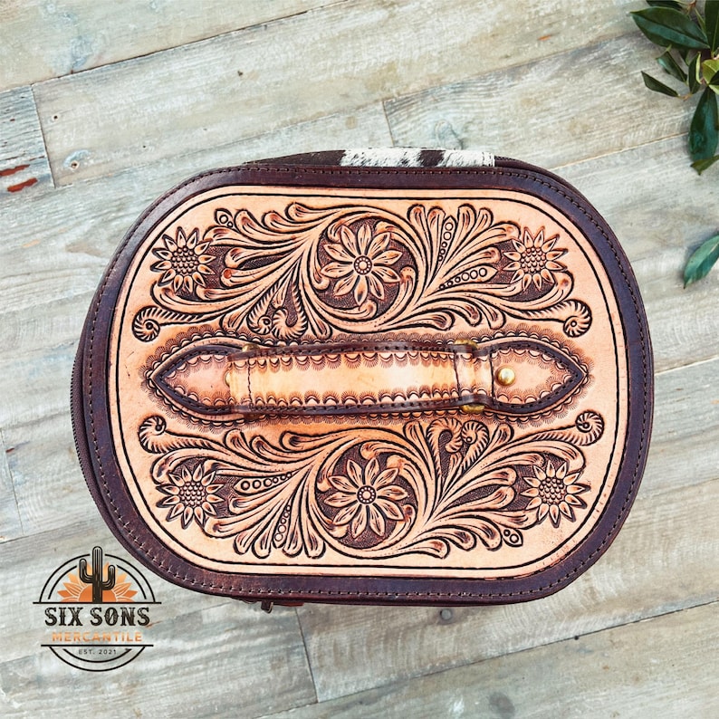 May include: A brown leather oval-shaped bag with a tooled floral design and a brown leather handle. The bag has a zipper closure and is made of high-quality leather.
