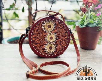 Puede incluir: Un bolso redondo de cuero marrón con un diseño floral. El bolso tiene una correa larga y una pequeña solapa redonda con un cierre de metal. El bolso está decorado con un patrón floral de girasoles y hojas.