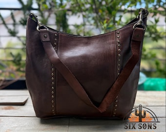 Puede incluir: Un bolso hobo de color marrón oscuro con filas verticales de pequeños remaches metálicos en los paneles frontales. El bolso incluye una correa ancha para el hombro sujeta con clips de latón antiguo y el sello SIX SONS MERCANTILE EST 2021.