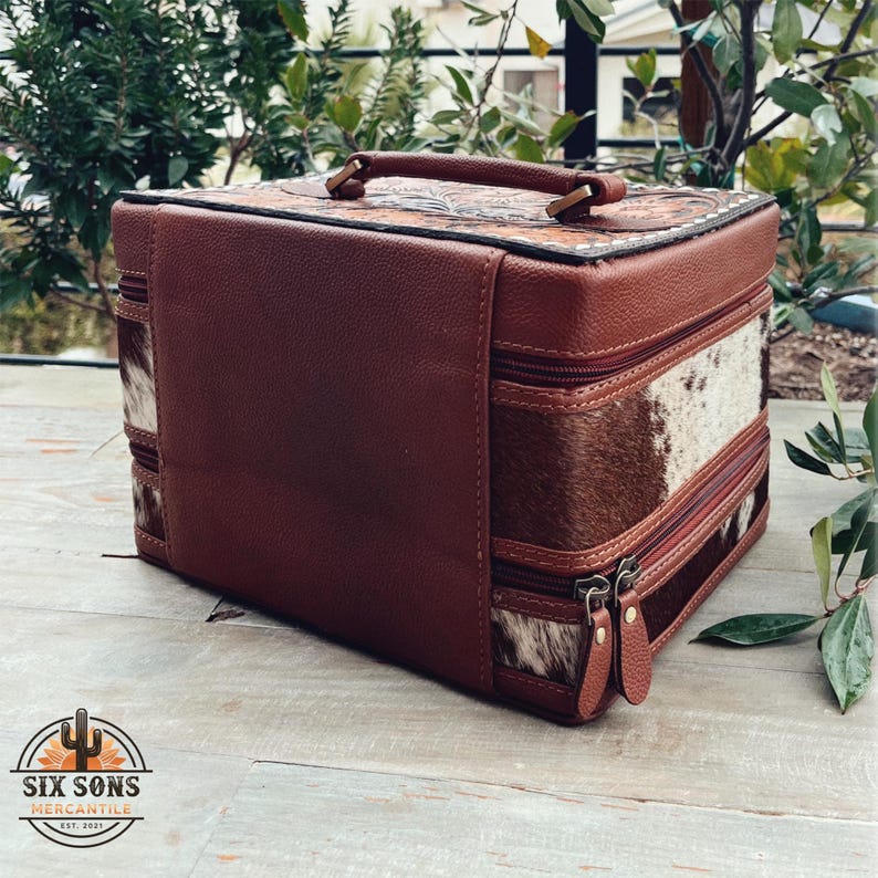 May include: A brown leather and cowhide makeup case with two zippers and a top handle. The case has a floral design on the top and is closed.