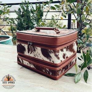 May include: A brown leather and cowhide two-tiered makeup case with a gold handle. The case has a floral design on the top and a cowhide pattern on the bottom.
