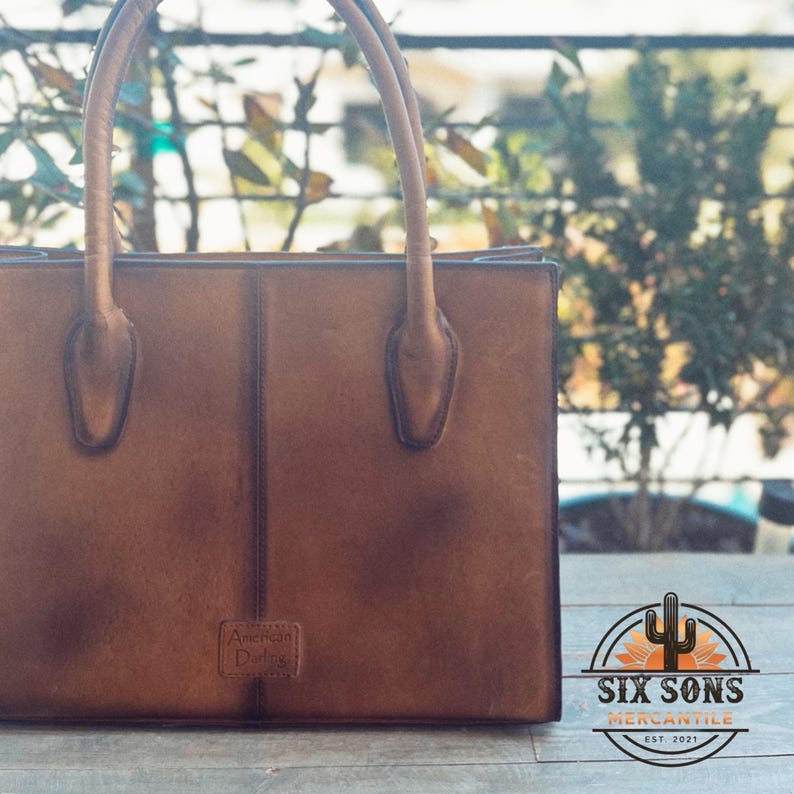 Puede incluir: Un bolso rectangular de cuero marr&oacute;n con dos costuras verticales, dos asas superiores redondeadas y un peque&ntilde;o parche en relieve en la parte inferior central que dice American Darling. El bolso tiene un acabado liso y mate.