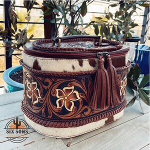 May include: Oval-shaped, brown leather cosmetic bag with a white and brown cowhide pattern. The bag has a zipper closure, a top handle, and two brown tassels. The bag is decorated with intricate floral carvings.