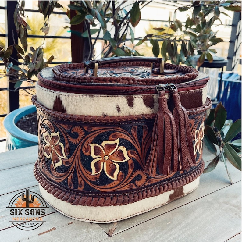 May include: Oval-shaped, brown leather cosmetic bag with a white and brown cowhide pattern. The bag has a zipper closure, a top handle, and two brown tassels. The bag is decorated with intricate floral carvings.