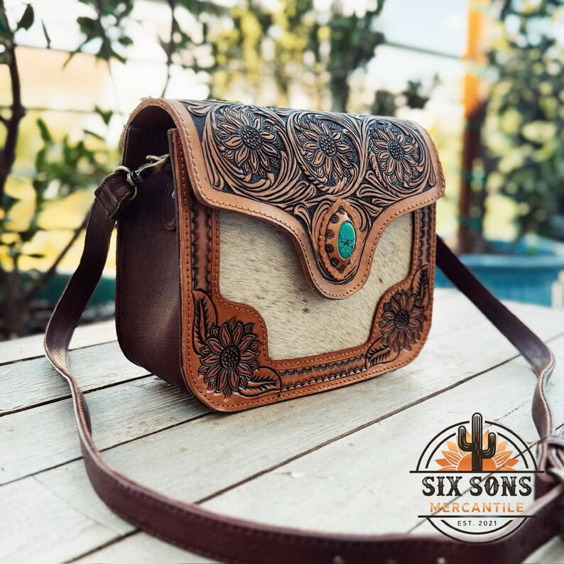 May include: A brown leather crossbody bag with a floral design and a turquoise stone clasp. The bag features a light-colored hair-on hide front panel and a long, adjustable strap. The bag has the text "SIX SONS MERCANTILE EST. 2021" on the bottom right.