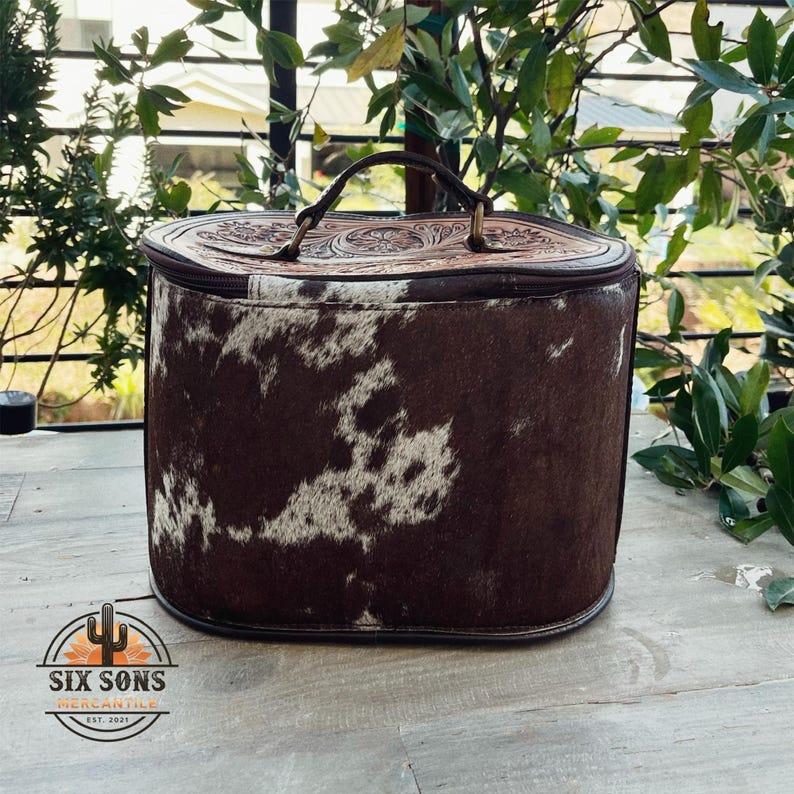 May include: A brown leather and cowhide travel bag with a top handle and a floral embossed design. The bag is closed with a zipper and has a brown leather trim.