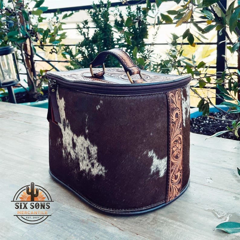 May include: A brown leather travel bag with a white and brown cowhide pattern. The bag has a top handle and a zipper closure. The bag is decorated with a floral design on the side.