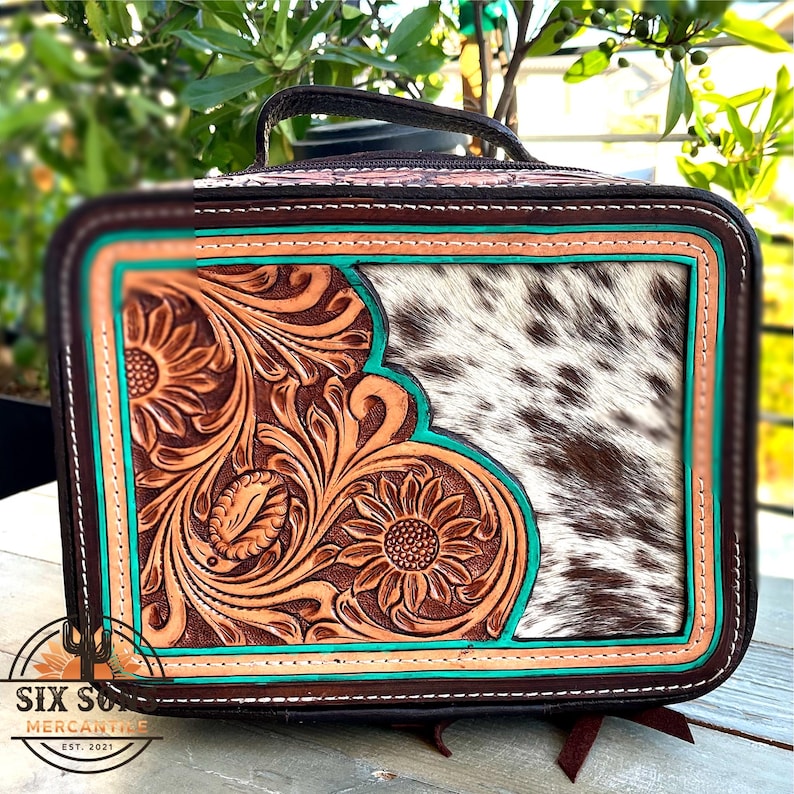 May include: A rectangular, brown leather bag with a handle, featuring a floral tooled design in tan and turquoise accents. The right side has a cowhide panel. The bag has the text "SIX SUNS MERCANTILE EST. 2021".
