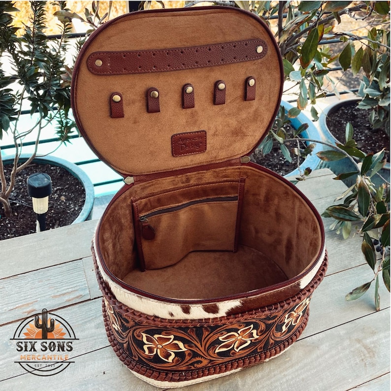 May include: A round, white and brown leather travel case with a floral design. The case has a brown leather interior with a zippered pocket and a row of small loops.