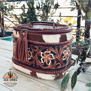May include: A brown and white oval-shaped leather bag with a floral design and a brown tassel. The bag has a brown leather handle and a zipper closure.