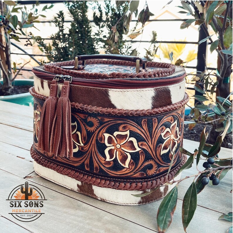 May include: A brown and white oval-shaped leather bag with a floral design and a brown tassel. The bag has a brown leather handle and a zipper closure.