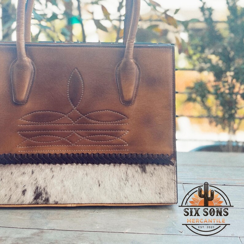 Puede incluir: Un bolso de cuero marr&oacute;n con un patr&oacute;n de costura decorativo en la parte superior, un borde trenzado oscuro y un panel de piel de vaca blanca y negra en la base. El bolso incluye dos asas de cuero y un logotipo que dice SIX SONS MERCANTILE EST. 2021.