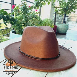 May include: A brown fedora hat with a black band. The hat has a wide brim and is made of felt. The hat is sitting on a white wooden surface. The text "Six Sons Mercantile Est. 2021" is printed on the brim of the hat.