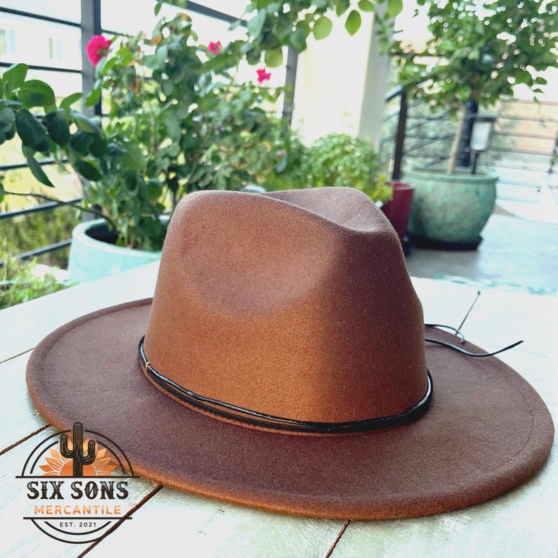 May include: A brown fedora hat with a black band. The hat has a wide brim and is made of felt. The hat is sitting on a white wooden surface. The text "Six Sons Mercantile Est. 2021" is printed on the brim of the hat.