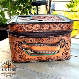 May include: A rectangular, brown leather bag with intricate floral and scroll tooling. The bag features a zipper closure, a handle, and turquoise accents. The logo "SIX SONS MERCANTILE" is visible on the bottom left.