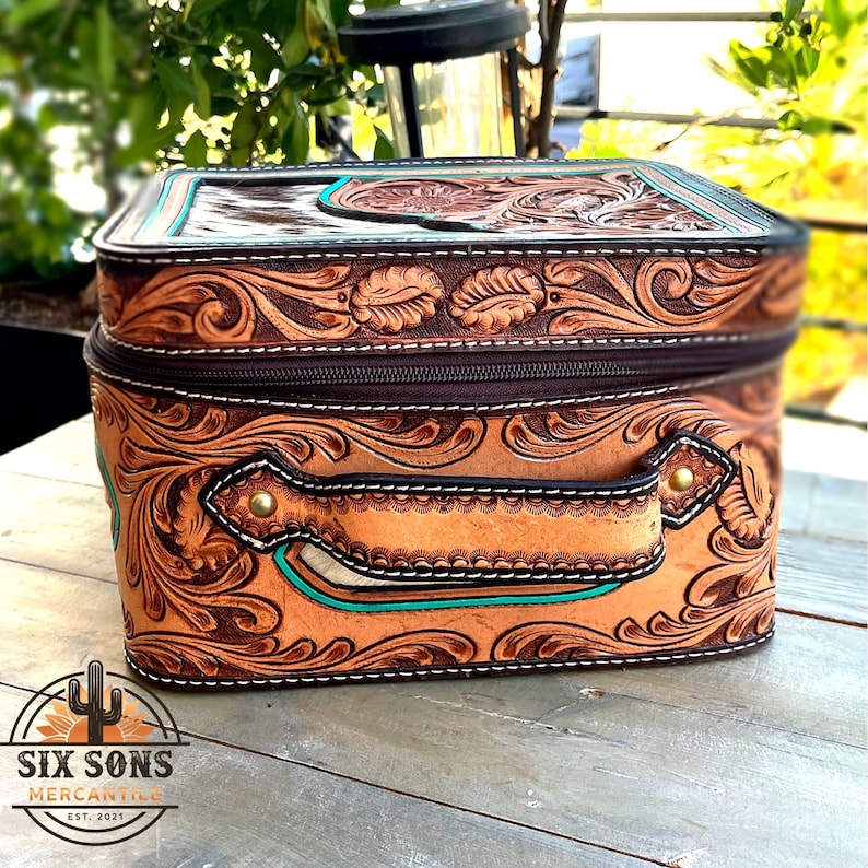 May include: A rectangular, brown leather bag with intricate floral and scroll tooling. The bag features a zipper closure, a handle, and turquoise accents. The logo "SIX SONS MERCANTILE" is visible on the bottom left.