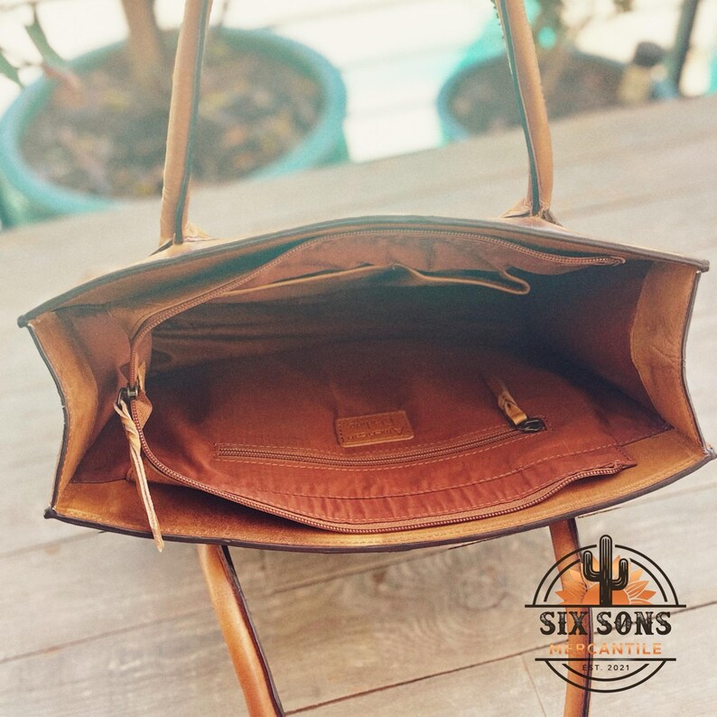 Puede incluir: Un bolso tote de cuero color canela con apertura superior y dos correas para el hombro. El interior est&aacute; forrado con tela marr&oacute;n y cuenta con un compartimento central con cremallera, un bolsillo lateral con cremallera y una etiqueta.