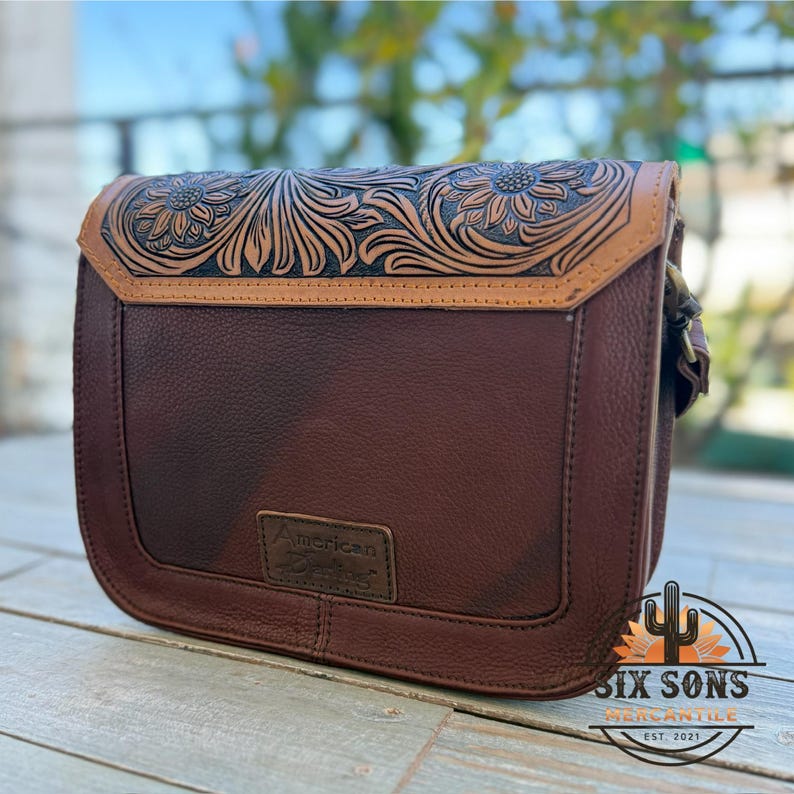 May include: A brown leather crossbody bag with a floral embossed design on the flap. The bag features a rectangular shape with a curved bottom and a buckle closure. The bag has a small rectangular tag that says "American Darling".