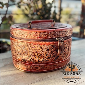 May include: A round, brown leather box with a tooled floral design. The box has a zipper closure and a handle on the top. The box is sitting on a wooden surface. The text "Six Sons Mercantile Est. 2021" is visible in the bottom right corner of the image.