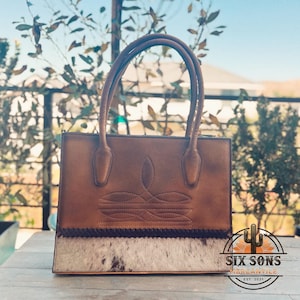 Puede incluir: Un bolso de cuero marr&oacute;n con dos asas, un patr&oacute;n decorativo cosido en la secci&oacute;n superior y un panel de piel de vaca texturizada en la base con un borde trenzado oscuro. El logotipo Six Sons Mercantile Est. 2021 es visible en la esquina.